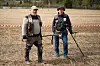 Two detectorists stand in a field with metal detectors and a red-and-white tape barrier behind them.