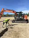 A field with an excavator and an archaeologist holding a metal detector.