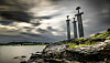 Three stone swords monument on a rocky shore beside Hafrsfjord under a cloudy sky.