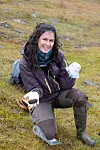 Researcher kneels on a grassy slope holding reindeer droppings in gloved hands.