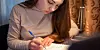 Teenage girl writing in a notebook beside a laptop and lamp at a desk.