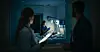 Hospital staff stand near a patient bed in a dark ward lit by monitors and medical lamps.