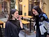 Two people smiling and talking outside a café with outdoor seating.