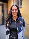 Smiling woman in glasses wearing a black and blue branded jacket in a narrow alley.