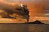 An ash plume rises from a volcano over the ocean beneath orange sunset clouds.