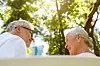 An older couple talking outdoors beneath trees in bright sunlight.