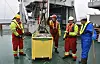 Crew members in hard hats and hi-vis clothing gather around a yellow equipment container on a ship deck.