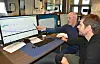 Two men examining stock charts on large computer screens in an office.