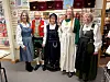 Five women in traditional costumes stand in front of shelves with textiles and goods.
