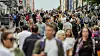 Crowded urban street with pedestrians walking in both directions between buildings.
