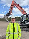 Man in hi-vis clothing stands beside an excavator on a dirt site.