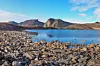 Rocky shoreline beside Lake Stuptjørna with calm water and mountains under a blue sky in Svalbard.
