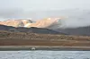 Small vehicle on a beach with water in the foreground and low mountains under cloud behind.