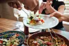 Hands serving food over a restaurant table with pizza, pasta, salad and drinks.