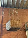 A wicker balloon basket hangs from the ceiling indoors in front of a red brick wall