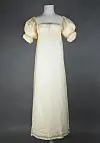 A light-coloured long dress with short puffed sleeves displayed on a mannequin against a dark background.