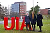 Three people posing beside large red UIA letters on a lawn with buildings behind them.