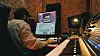 Person working alone at a computer in a home studio with a keyboard and music software on screen.