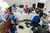 Cluttered bedroom with a bed covered in clothes, scattered laundry, a desk, and a TV.