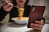 A young woman scrolls on her phone while eating pasta at a dinner table.