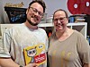 To personer smiler og holder en fargerik barnbok innendørs foran en hylle.