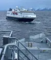 Passenger ferry on rough water during a safety exercise, viewed from another vessel with deck equipment in the foreground.