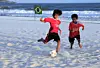 Two boys play football on a beach with the ocean as a backdrop. A Brazilian flag is set up on the edge of where the boys are playing.