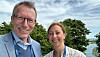 Portrait of Bjørn Naume and Hege Oma Ohnstad. They are standing outdoors with a little wind in their hair, in the background trees can be seen, water can be glimpsed, and a blue sky with scattered clouds.