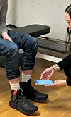 Person seated in black boots and rolled-up jeans while another holds a smartphone in a plain room.