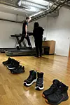 Person walking on a treadmill in a lab while another person monitors; several black shoes are lined up in the foreground.