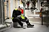 Worker crouching in an industrial plant, inspecting a black safety boot beside pipes and valves.