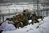 Soldiers in camouflage lie prone in snow during a winter training exercise with rifles.
