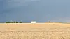 A small white house stands on a hill above a golden field under a dark sky in rural Iowa.