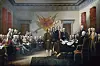 Officials and delegates stand and sit around a table in a historic chamber during a formal document signing.