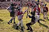 Reenactors in period military uniforms stage a mock battle on a grassy field.