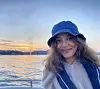 Portrait of a smiling Hijab Fatima Ali. She is on a boat, wearing a life jacket and a canvas boating hat. In the background, the sea and a sunset are visible.