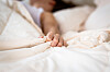 A hand grips a white bedsheet. The upper body of a woman can be seen. She has dark hair and is wearing a white top. The image is meant to depict her having an orgasm.