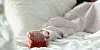 A hand holding a glass of a red drink while sitting in a bed with white sheets.