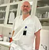 A man in a white medical uniform stands in a bright clinic room with shelves, bottles, and a sink in the background.