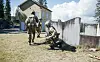 Two soldiers in tactical gear train beside a concrete wall during an outdoor exercise.