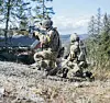 Soldiers in camouflage gear conduct a woodland training exercise with rifles and tactical equipment.