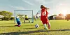 Children training in football on a grassy pitch with a goal and ball at sunset.