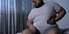 Overweight man sitting near a window, holding his stomach.