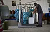 A worker fills a tank with olive oil at an olive mill equipped with blue machinery.