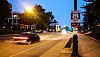 Evening traffic moves past a Route 66 sign on a city street lined with buildings and trees.