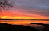 Orange and pink sunset over a calm lake with dark shoreline silhouettes and leafless trees.