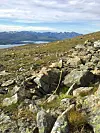 A rocky mountainside with grass, a lake, and mountains in the background.