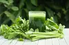 A glass of green celery juice in front of fresh celery on a light-coloured table.
