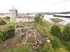 Avaldsnes Church in the background, with the wall of the old medieval royal estate visible in the foreground. Excavation is underway. To the right, the fjord and a ship can be seen.