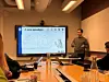 A man presents mobility research beside a large screen in a conference room with attendees seated at a table.